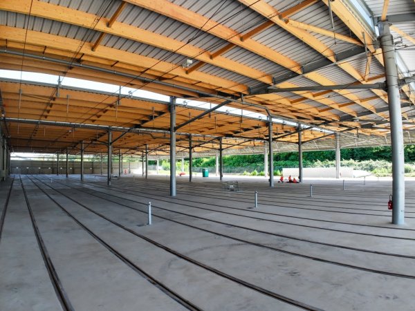 Intérieur du centre de maintenance de tramway de Saint-Fons, avec plusieurs voies ferrées parallèles intégrées dans le sol en béton. La structure est composée de nombreux poteaux métalliques soutenant une charpente en bois clair et une toiture industrielle. À l'arrière-plan, de larges ouvertures laissent entrer la lumière naturelle et donnent sur une zone végétalisée. (Crédit : Éric Soudan - Alpaca Productions)