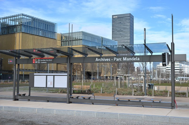 Station de la ligne TB12 "Archives – Parc Mandela", avec une structure en métal et un toit en verre. À l'arrière, on voit des bâtiments contemporains aux façades vitrées, un espace vert avec des arbres et des bancs, ainsi que le gratte-ciel To-Lyon au loin. Le décor est urbain et dégagé sous un ciel partiellement bleu. Crédit photo : Éric Soudan - Alpaca Productions