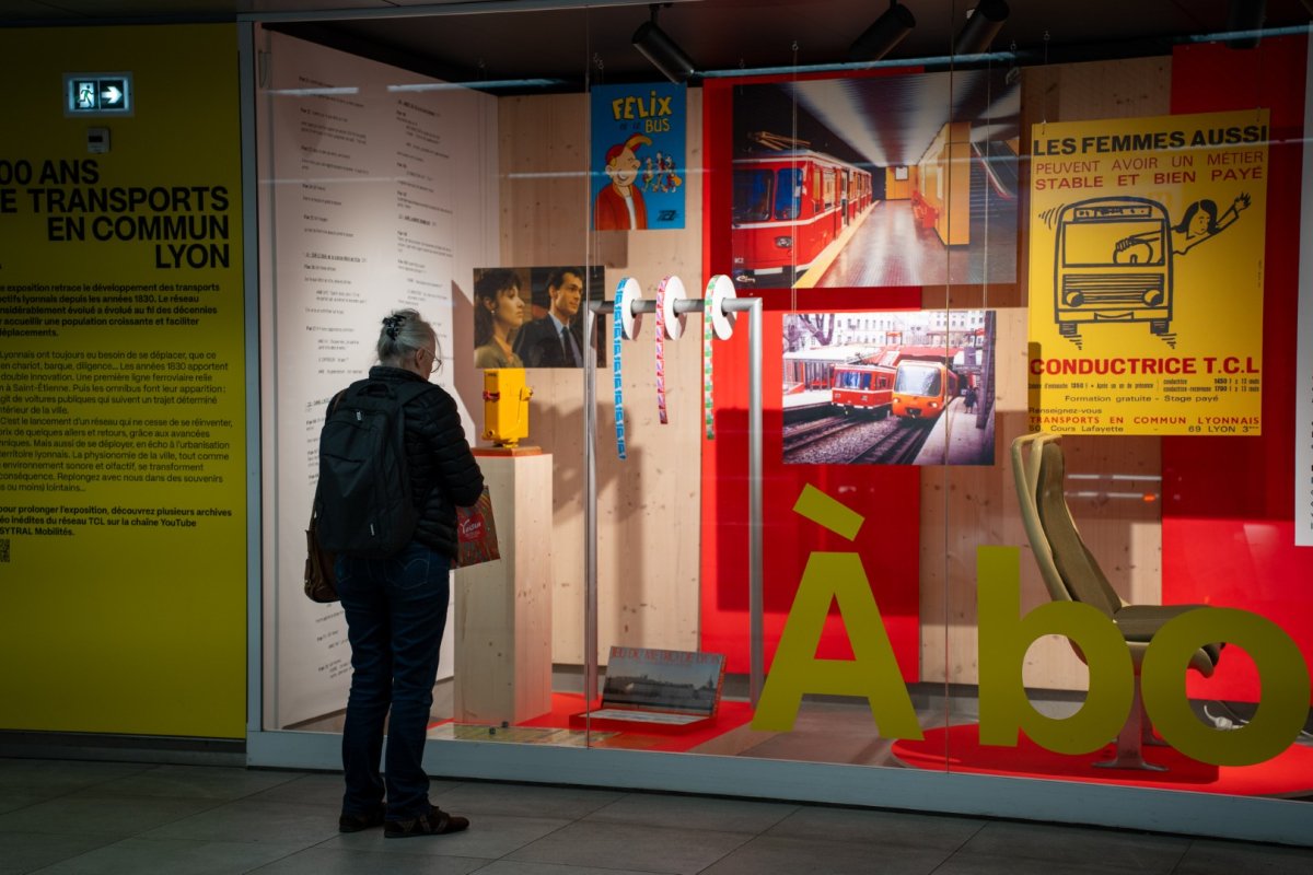 Vitrine d'exposition sur l'histoire des transports en commun lyonnais : une personne vue de dos observe des panneaux colorés présentant photos de tramways et métros anciens et récents, affiches historiques, textes explicatifs et objets liés au réseau TCL, dans un espace intérieur éclairé de la station de métro Part-Dieu.
