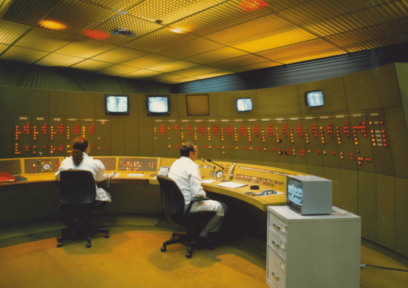 Photographie du poste de contrôle centralisé du métro, vers 1980 : deux opérateurs sont assis à un bureau de forme arrondie, face à un mur parsemé de plusieurs écrans et de diodes lumineuses