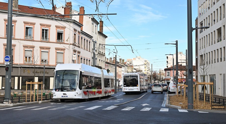 Deux trolleybus articulés circulent sur une rue dans le secteur Maisons Neuves, bordée d’immeubles anciens aux façades claires et de bâtiments plus modernes. Des lignes aériennes de contact et des lampadaires sont visibles en arrière‑plan. La scène montre une portion de voirie récemment aménagée avec trottoirs, traversées piétonnes et végétalisation. Crédit photo : Éric Soudan - Alpaca Productions