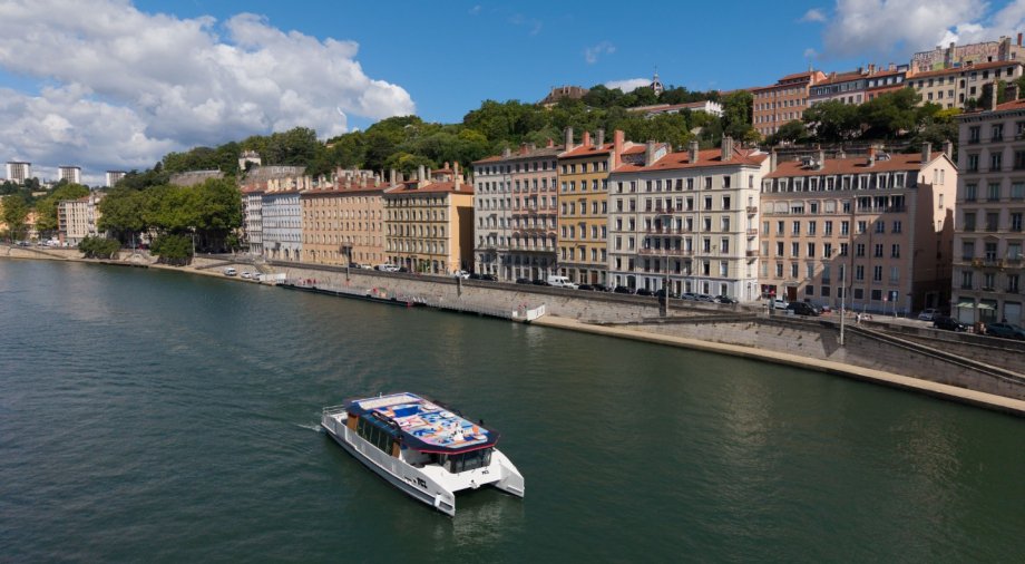 Navette fluviale du réseau TCL naviguant sur la Saône, le long des façades du Vieux Lyon sous un ciel ensoleillé (crédit : Éric Soudan - Alpaca Productions)