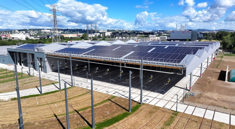 Vue aérienne du nouveau centre de maintenance de Saint-Fons. Sous la halle ouverte, plusieurs voies ferrées parallèles sont visibles, équipées de lignes aériennes et de poteaux. Crédit : Éric Soudan - Alpaca Productions