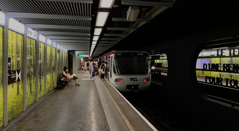 Station de métro Gratte-Ciel rénovée sur la ligne A. Une rame se situe le long du quai, avec des revêtements sombres, éclairage linéaire au plafond et bande de guidage tactile au sol. Ses parois sont décorées d'un motif de forêt aux tons verts. Plusieurs voyageurs attendent ou montent à bord.
